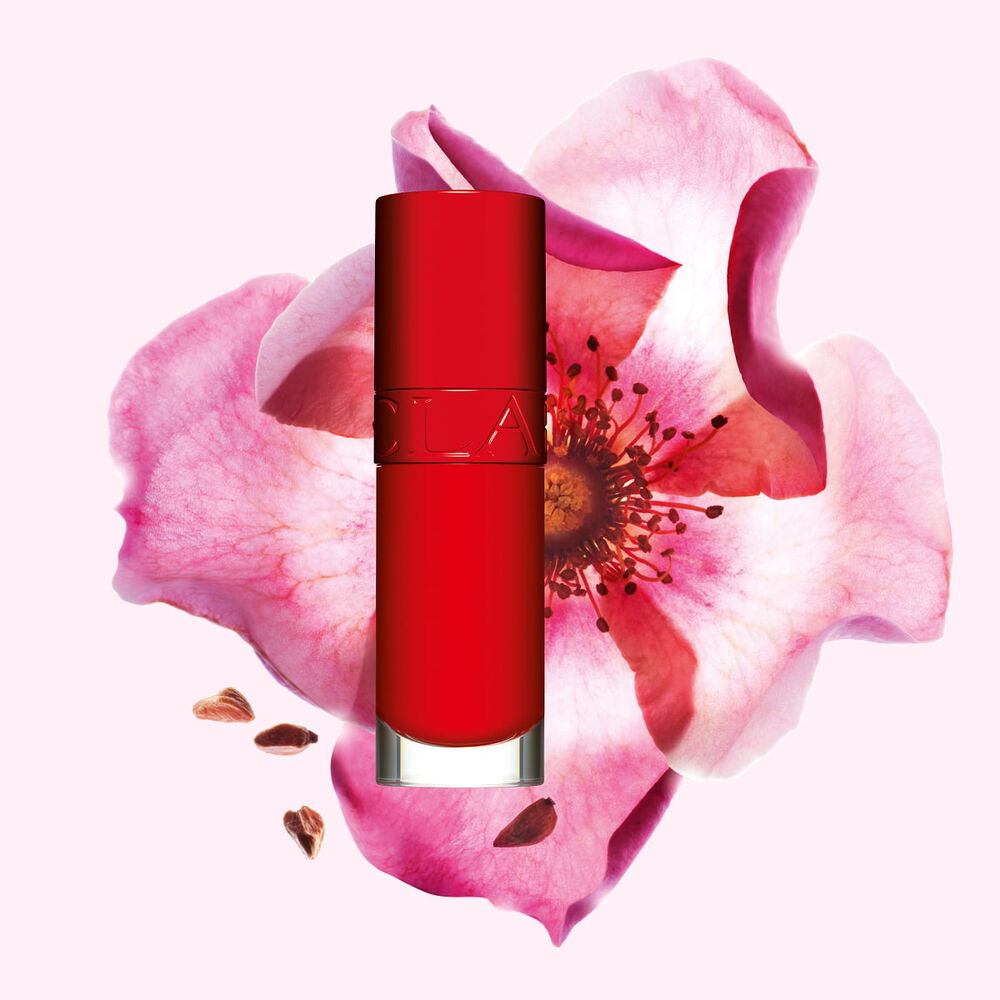 Packshot av Clarins Lip Comfort Oil i Iconic Red, plassert ved siden av en nyperoseblomst p&aring; en svart-r&oslash;d bakgrunn.