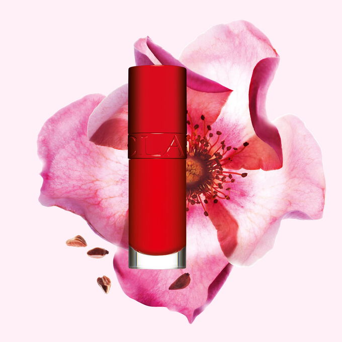 Packshot av Clarins Lip Comfort Oil i Iconic Red, plassert ved siden av en nyperoseblomst p&aring; en svart-r&oslash;d bakgrunn.
