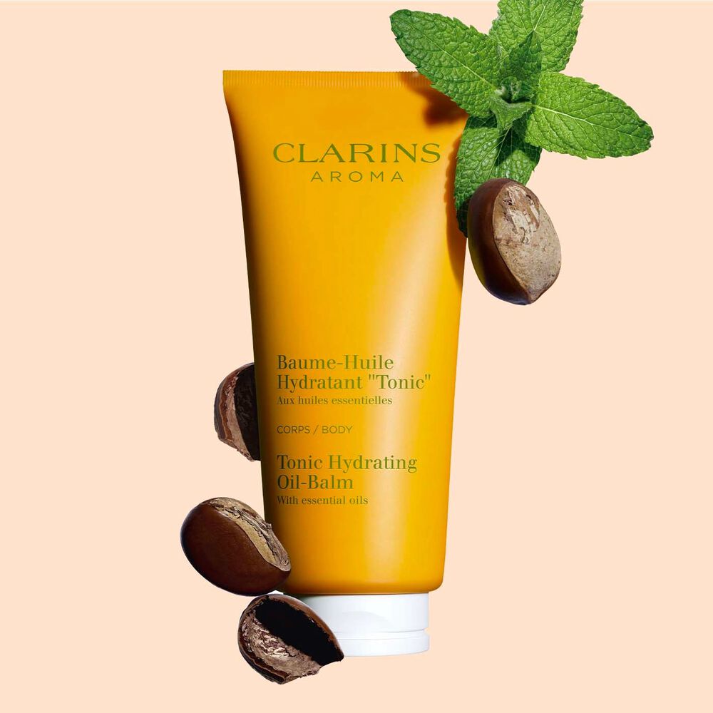 Pakkebilde av en tube med Clarins Tonic Hydrating Oil-Balm, i nærheten av mynteblader og sheafrø mot en ferskenfarget bakgrunn.