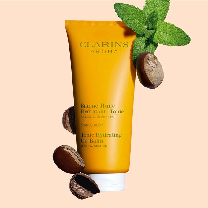Pakkebilde av en tube med Clarins Tonic Hydrating Oil-Balm, i nærheten av mynteblader og sheafrø mot en ferskenfarget bakgrunn.