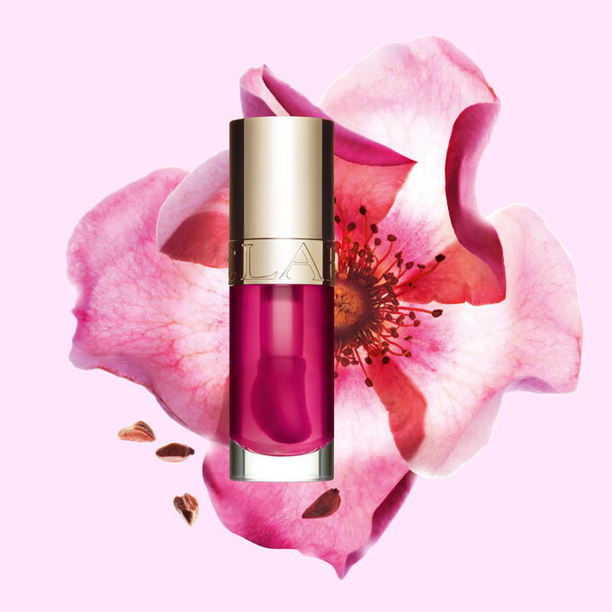 Packshot av Clarins Lip Comfort Oil i Raspberry, plassert ved siden av en nyperose p&aring; en lys ferskenfarget bakgrunn.