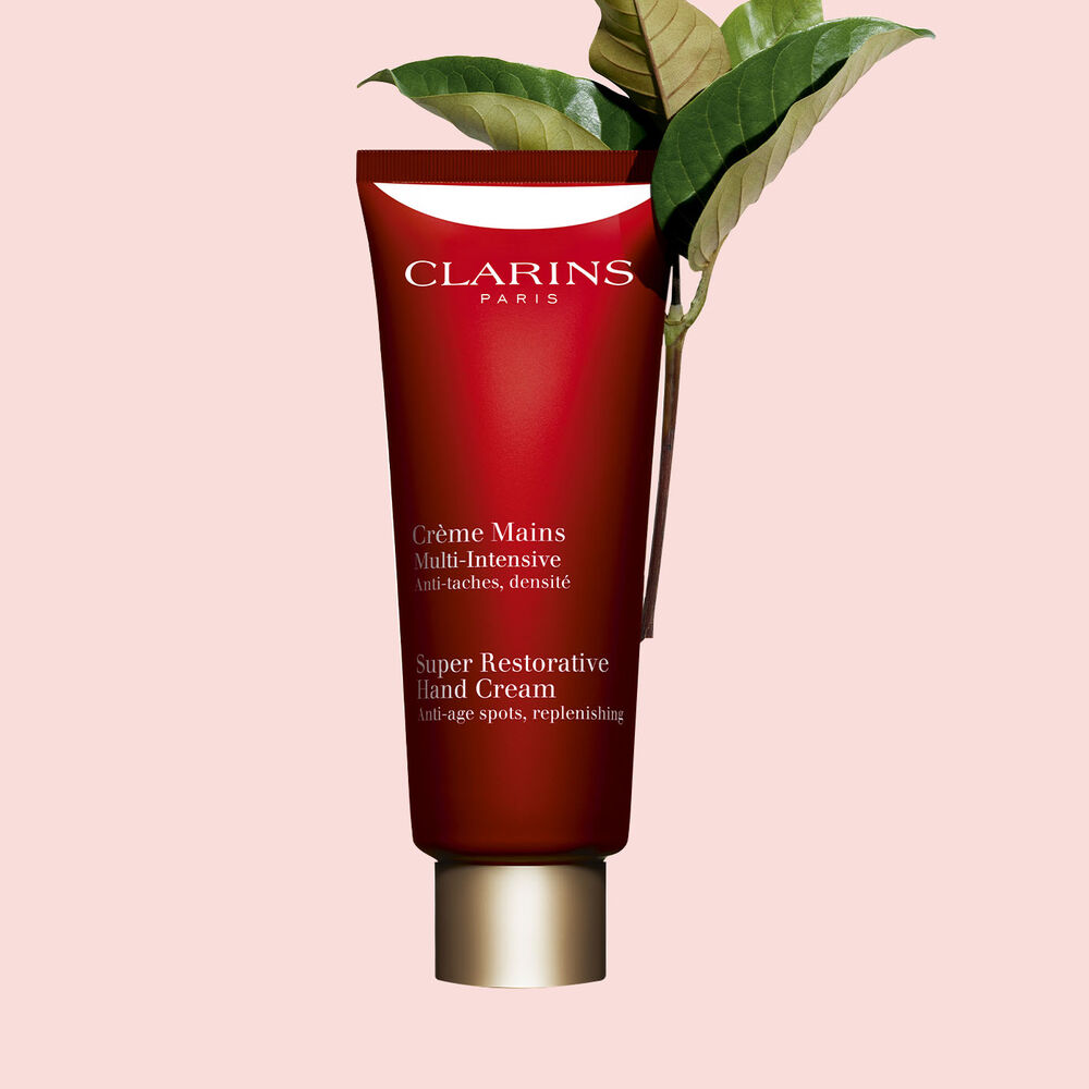 Pakkebilde av en flaske med Clarins Super Restorative Hand Cream i nærheten av noen harunganablader mot en rosa bakgrunn.