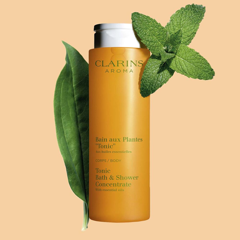 Pakkebilde av Clarins Tonic Bath and Shower Concentrate gel, i nærheten av mynte- og rosmarinblader mot en oransje bakgrunn.