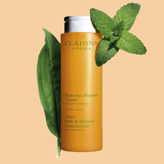 Pakkebilde av Clarins Tonic Bath and Shower Concentrate gel, i nærheten av mynte- og rosmarinblader mot en oransje bakgrunn.