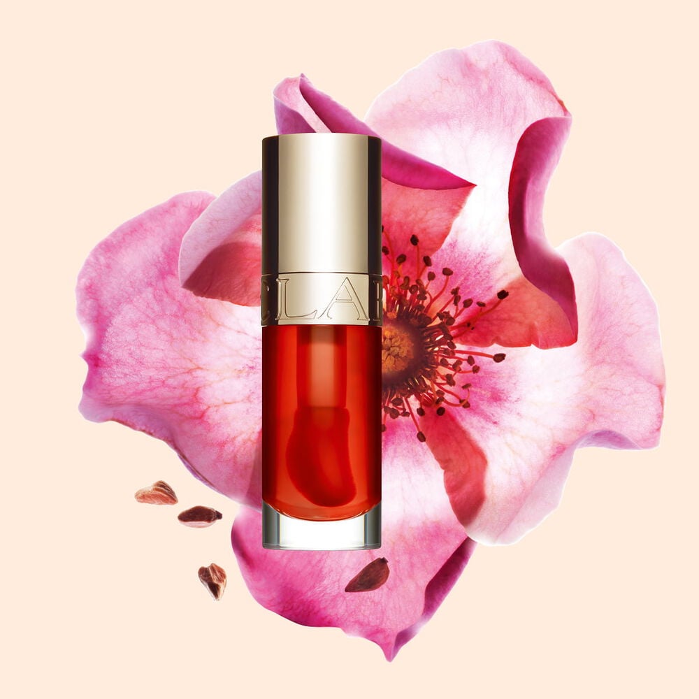 Packshot av Clarins Lip Comfort Oil i Apricot, plassert ved siden av en nyperose p&aring; en lys oransje ferskenfarget bakgrunn.