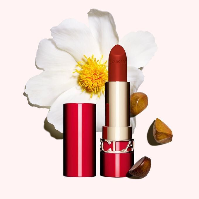 Packshot av Clarins matt leppestift Joli Rouge 782 V, ved siden av kamelia-blomst og sheafrø på rosa bakgrunn.