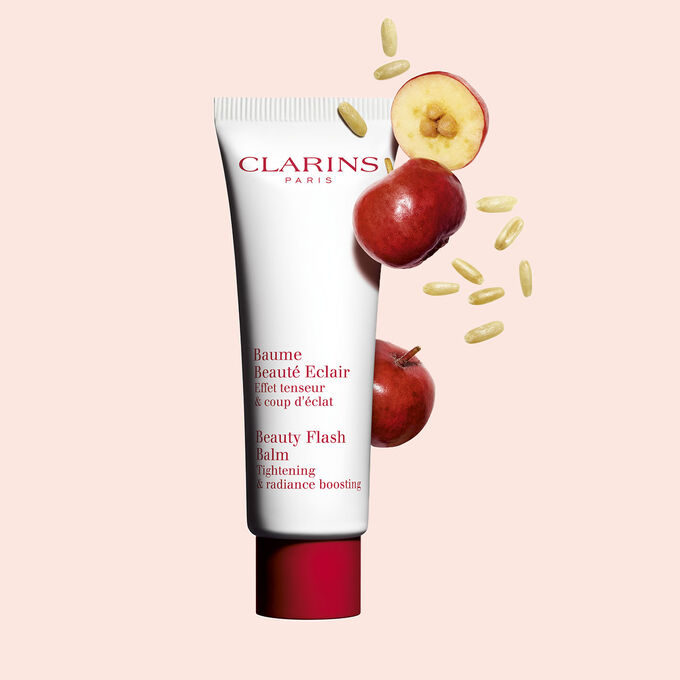 Pakkebilde av en tube med Clarins Beauty Flash Balm i nærheten av acerolafrø mot en ferskenfarget bakgrunn.