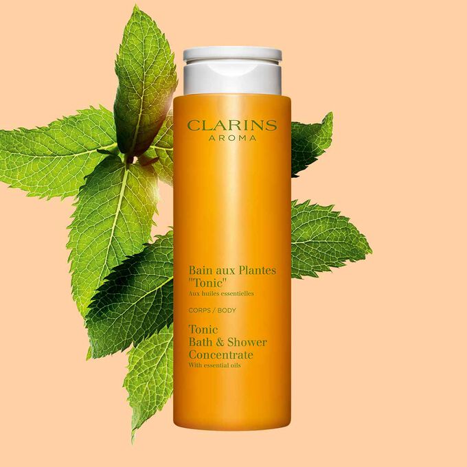 Pakkebilde av Clarins Tonic Bath and Shower Concentrate gel, i nærheten av mynte- og rosmarinblader mot en oransje bakgrunn.