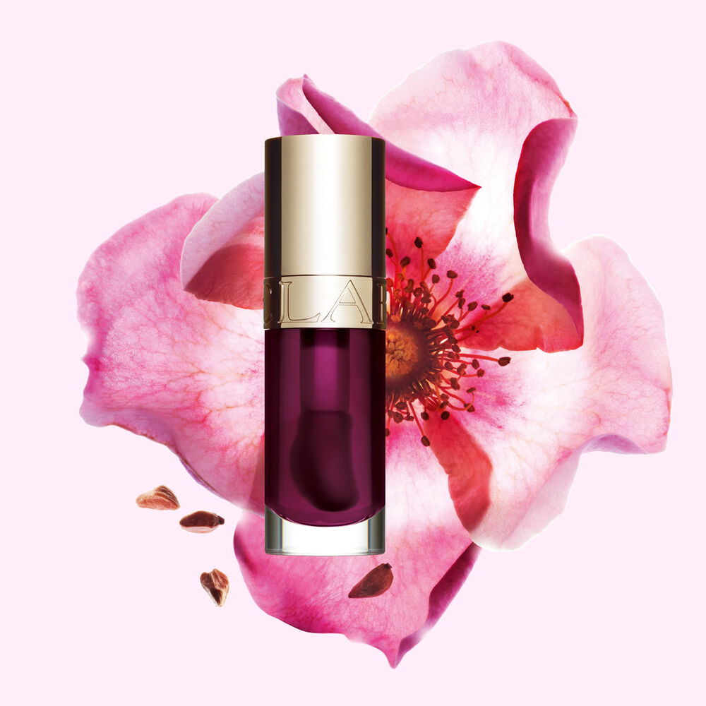 Packshot av Clarins Lip Comfort Oil i Plum, plassert ved siden av en nyperose p&aring; en lys rosa bakgrunn.