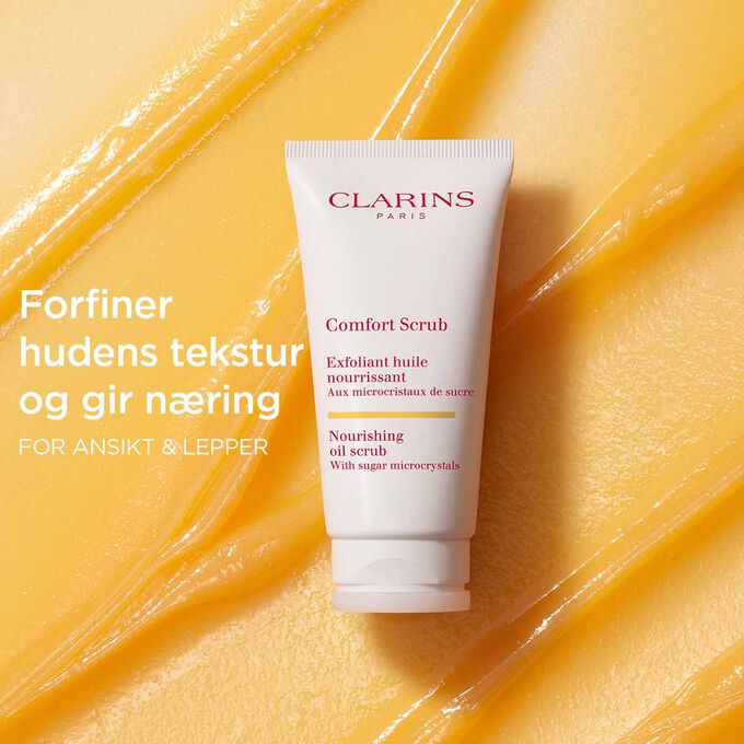Clarins Comfort Scrub for ansikt og lepper p&aring; en oransje, teksturert bakgrunn som fremhever dens forfinende og n&aelig;rende effekt.