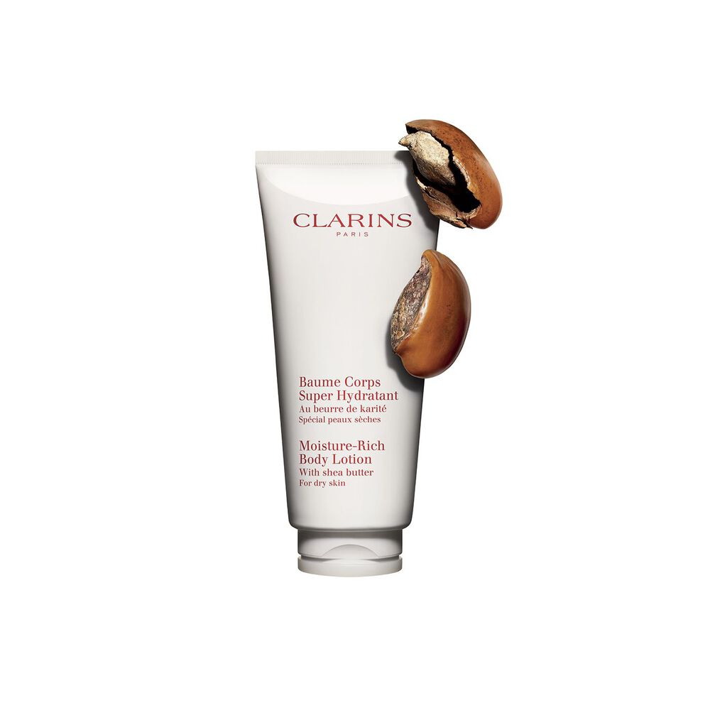 Pakkebilde av en tube med Clarins Moisture-Rich Body Lotion fra Clarins i nærheten av noen sheanøtter mot en hvit bakgrunn.